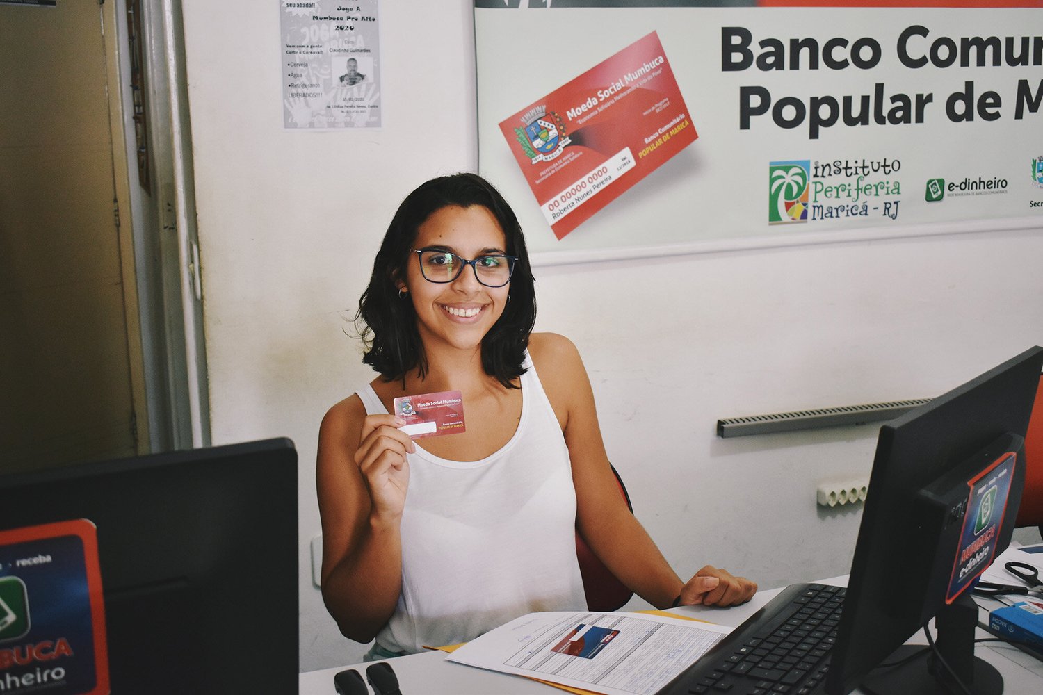 A woman sitting at a desk holding a red card, smiling. She is in an office with computers and a sign on the wall related to "Banco Comunitário Popular de Maricá."