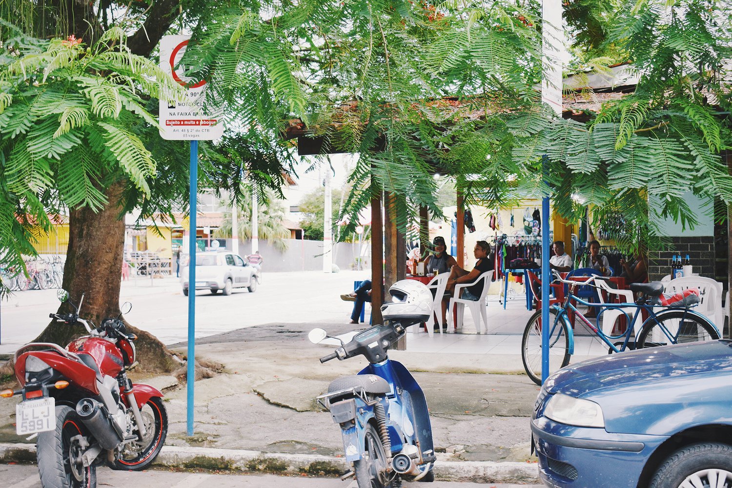 Outdoor cafe with people sitting on plastic chairs at tables under trees. Motorcycles and a car are parked nearby. A sign is visible behind a tree.